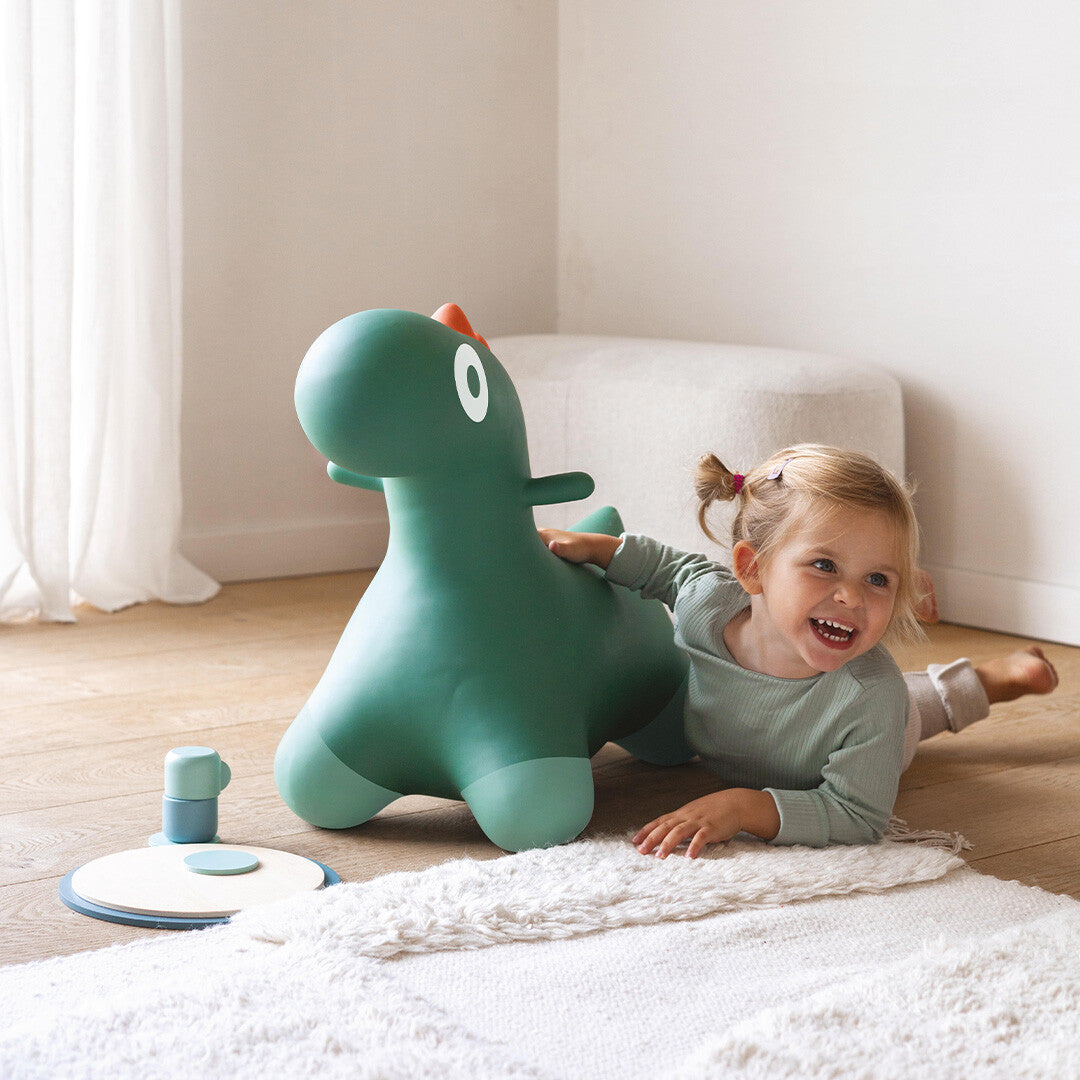 Een lachend kind ligt op de grond naast het Quut groene dinosaurus skippydier.