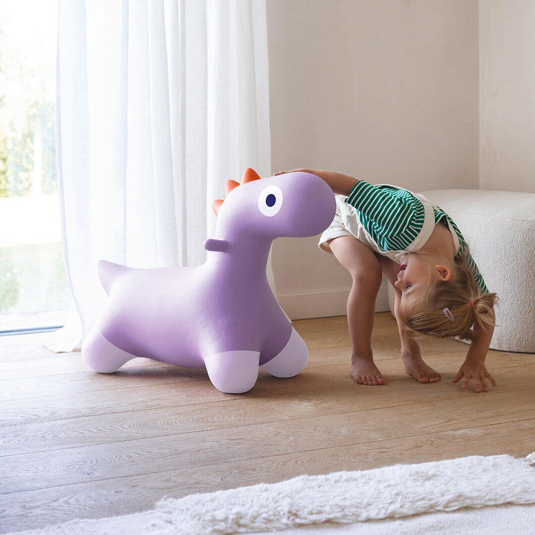 Een meisje buigt voorover naast het Quut paarse dinosaurusskippydier in een lichte kamer.