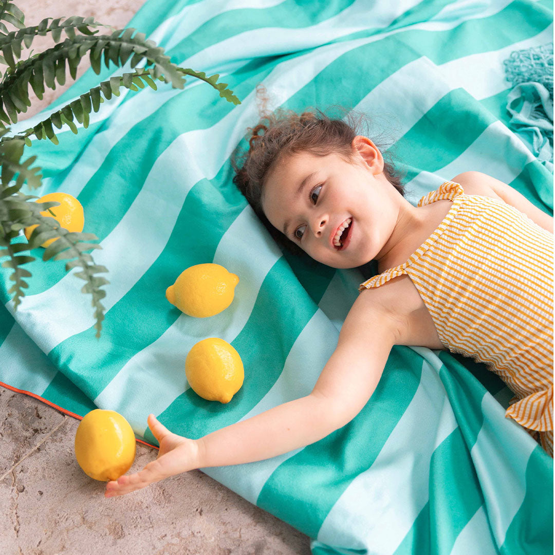 Een lachend meisje ligt op een gestreepte quut strandhanddoek in groen en lichtblauw.