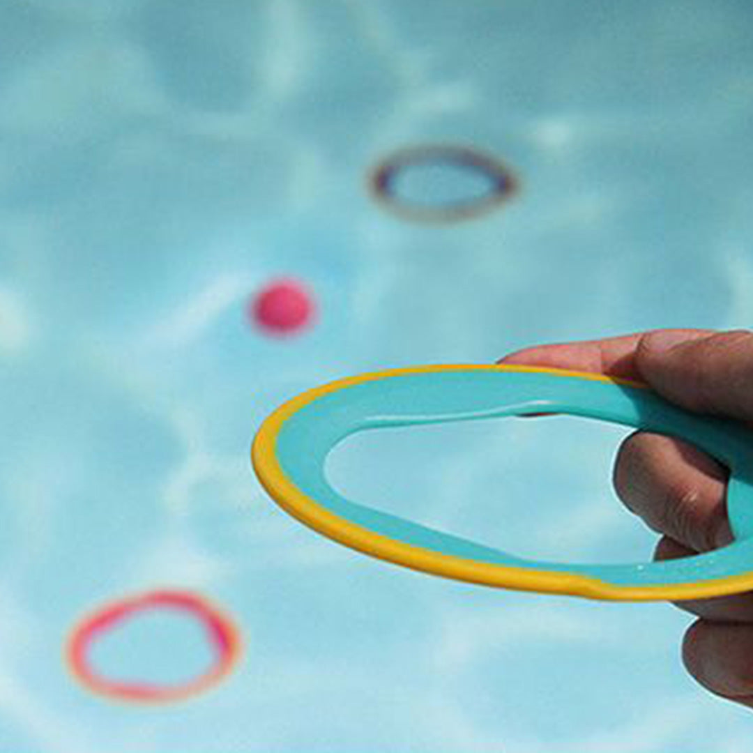 Een hand houdt een Quut Ringo ring vast in het water, met andere ringen en een bal drijvend op de achtergrond.