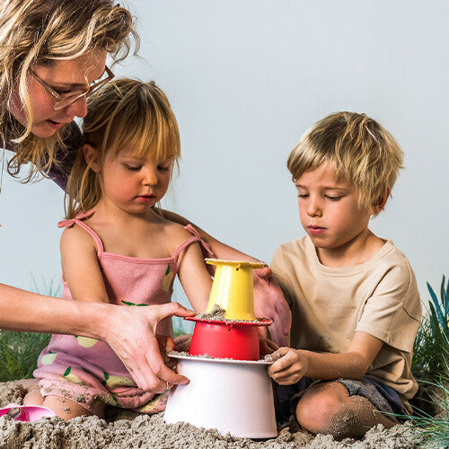 Een volwassene en twee kinderen stapelen de roze, rode en gele quut zandstapeltoren onderdelen in het zand.