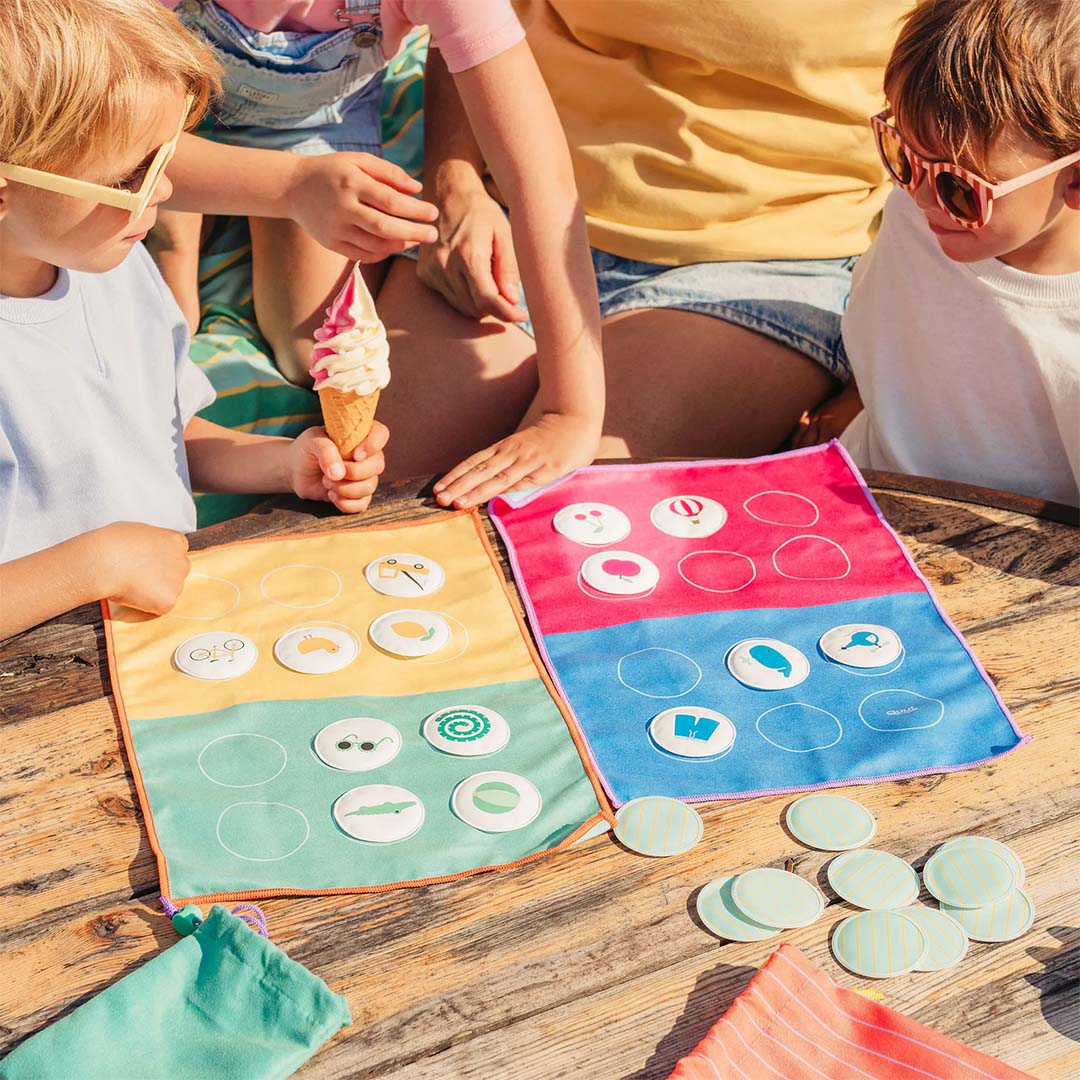 Kinderen spelen het Quut reisspel bingo met twee kleurrijke stoffen speelborden en fiches op een houten tafel.