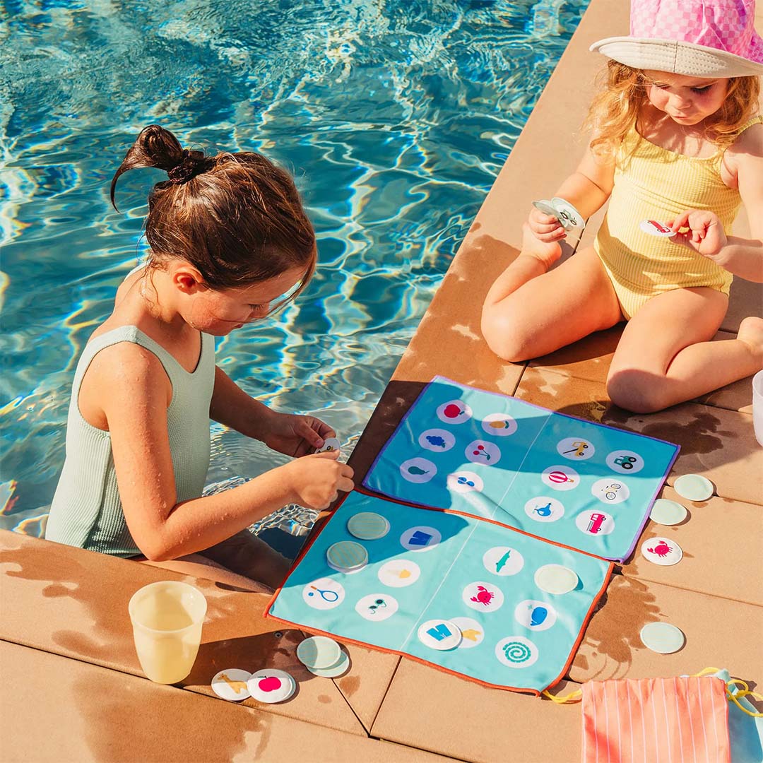 Twee meisjes spelen het Quut reisspel bingo met een blauw stoffen speelbord en fiches bij een zwembad.