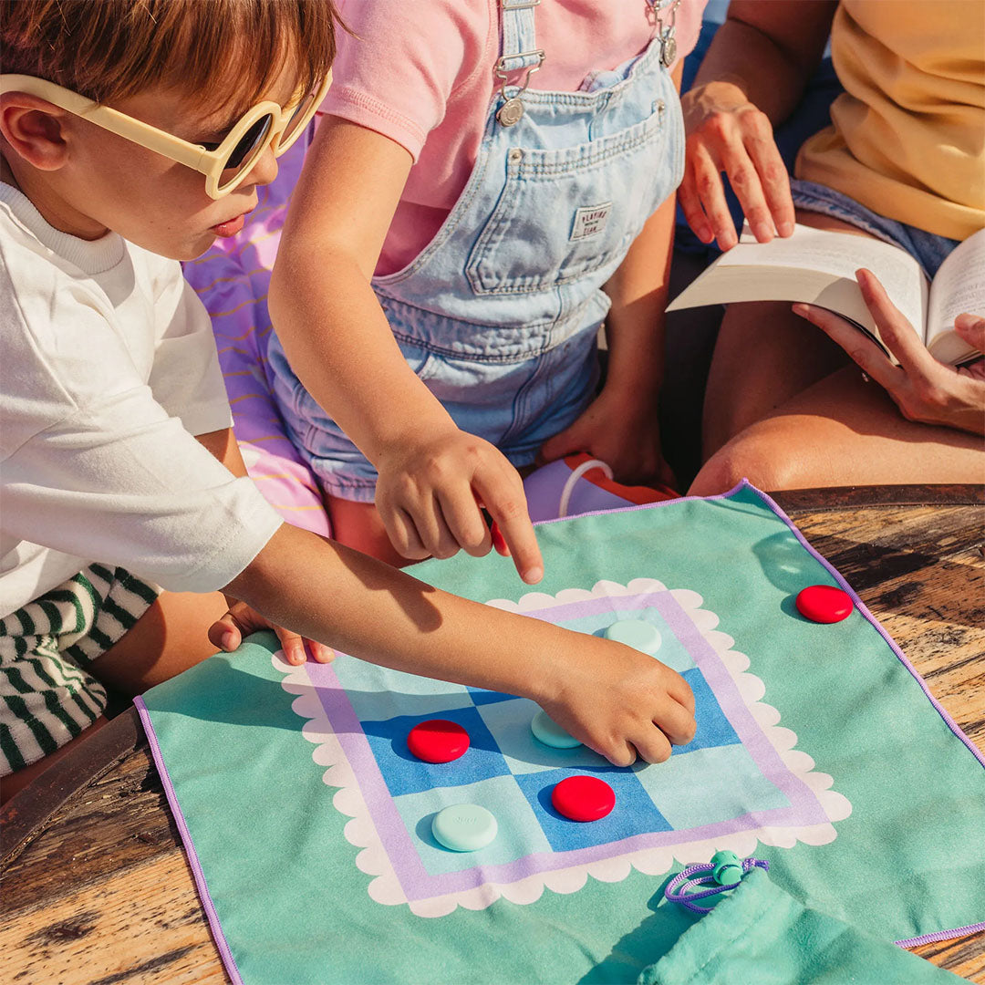 Kinderen spelen het quut reisspel boter kaas en eieren op een teal stoffen speelmat met rode en lichtblauwe speelstukken.
