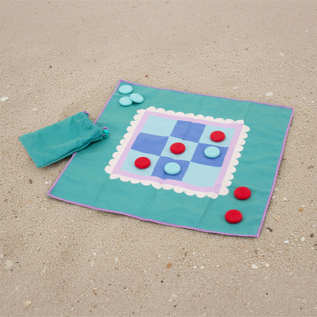 Het quut reisspel boter kaas en eieren op een teal stoffen speelmat met rode en lichtblauwe speelstukken, liggend op zand.