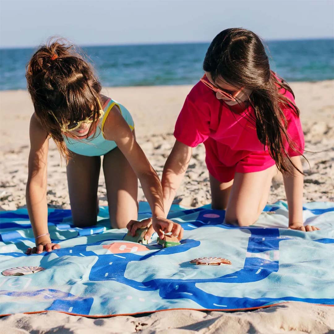 Twee meisjes spelen met speelgoedauto's op de quut speelhanddoek met wegenpatroon op een zandstrand.
