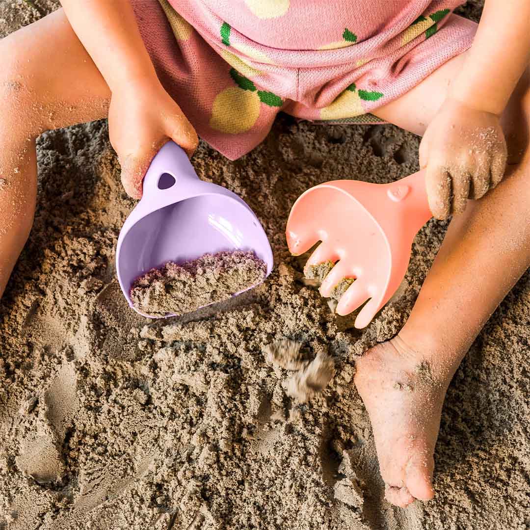 Een kind speelt met de Quut paarse schep en oranje hark in het zand.