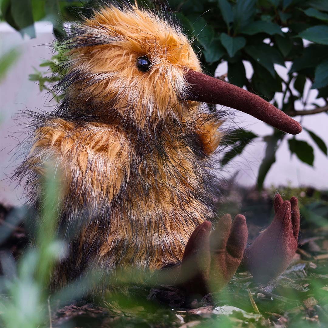 Rappa pluchen kiwi knuffelvogel zittend in groen struikgewas, schuin van voren gefotografeerd.
