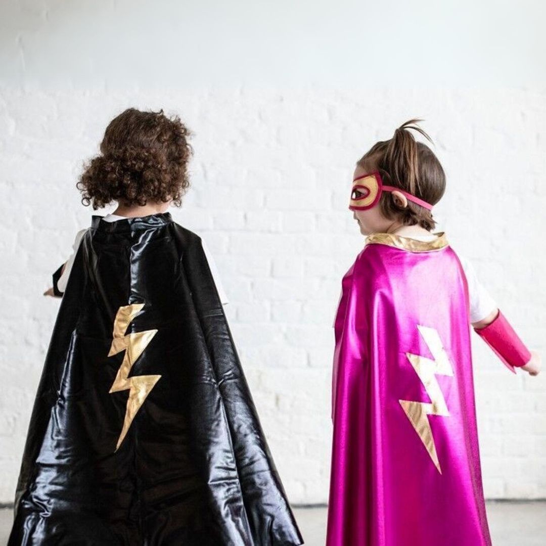Twee kinderen van achteren met superheldencapes, een zwarte en een roze, beide met een gouden bliksemschicht.
