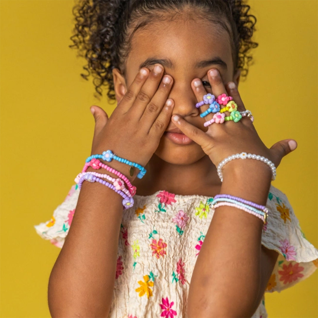 Meisje met donkere krullen bedekt haar ogen en draagt diverse kleurrijke kinderarmbanden en ringen.