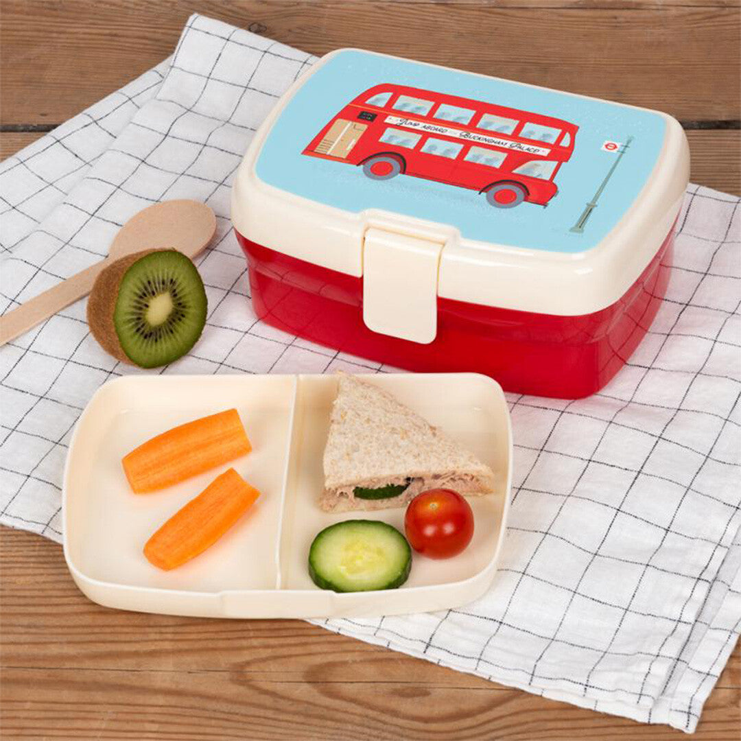 Een open Rex London broodtrommel met Routemaster bus-thema, gevuld met groenten en een sandwich, op een houten tafel met een theedoek.