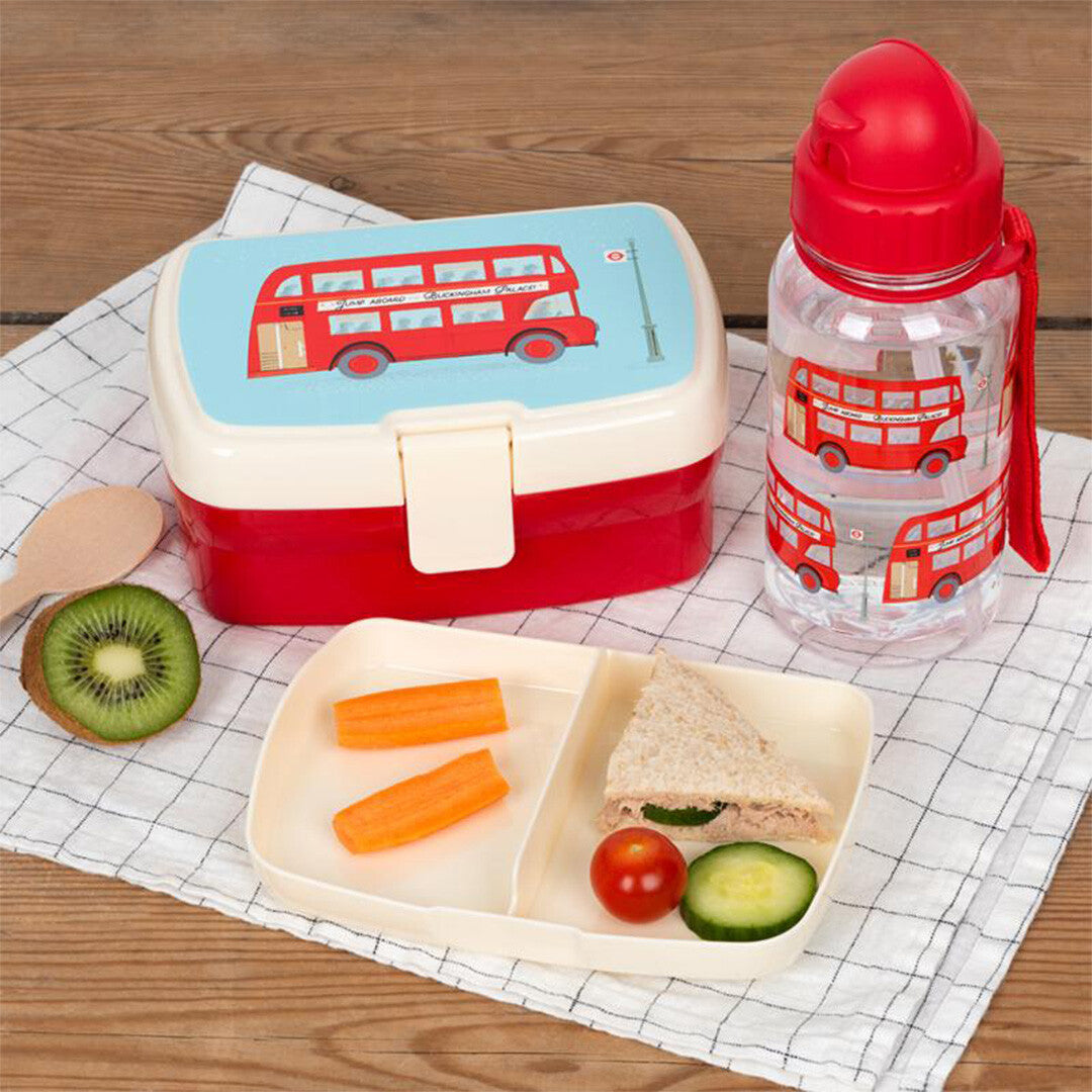 Een Rex London broodtrommel en bijpassende drinkfles met Routemaster bus-thema, gevuld met lunch en fruit, op een houten tafel.
