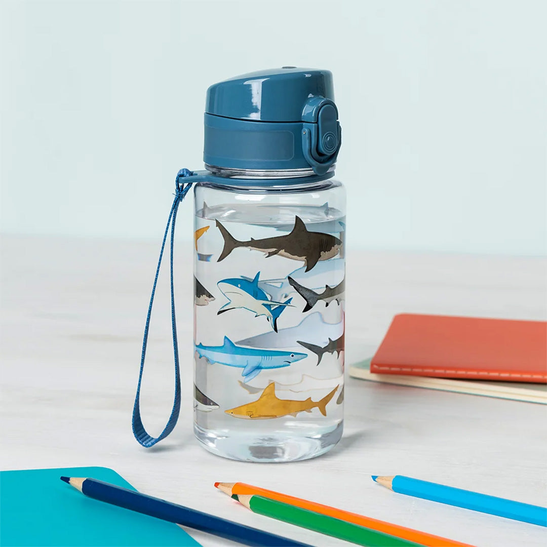 Rex London kinder drinkfles met haaienpatroon, gevuld met water, tussen kleurpotloden en notitieboekjes.