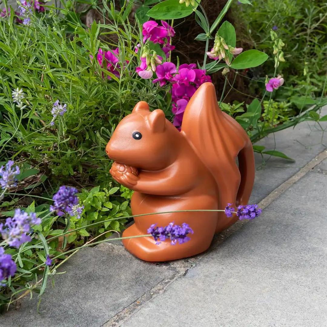 Bruine plastic eekhoorn gieter staat op een terras tussen paarse bloemen.