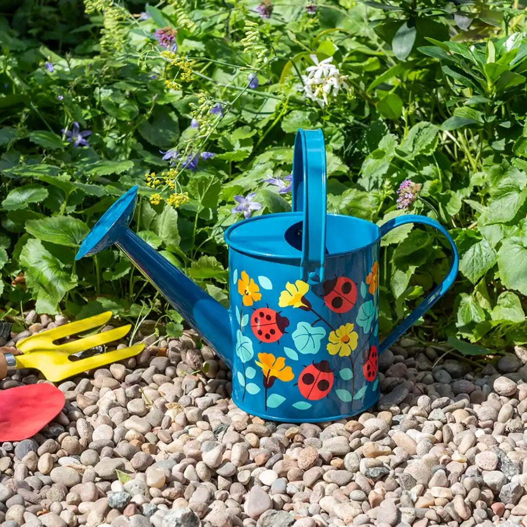 Rex London blauwe gieter met lieveheersbeestjes en bloemenpatroon op grind in een tuin, met tuingereedschap ernaast.