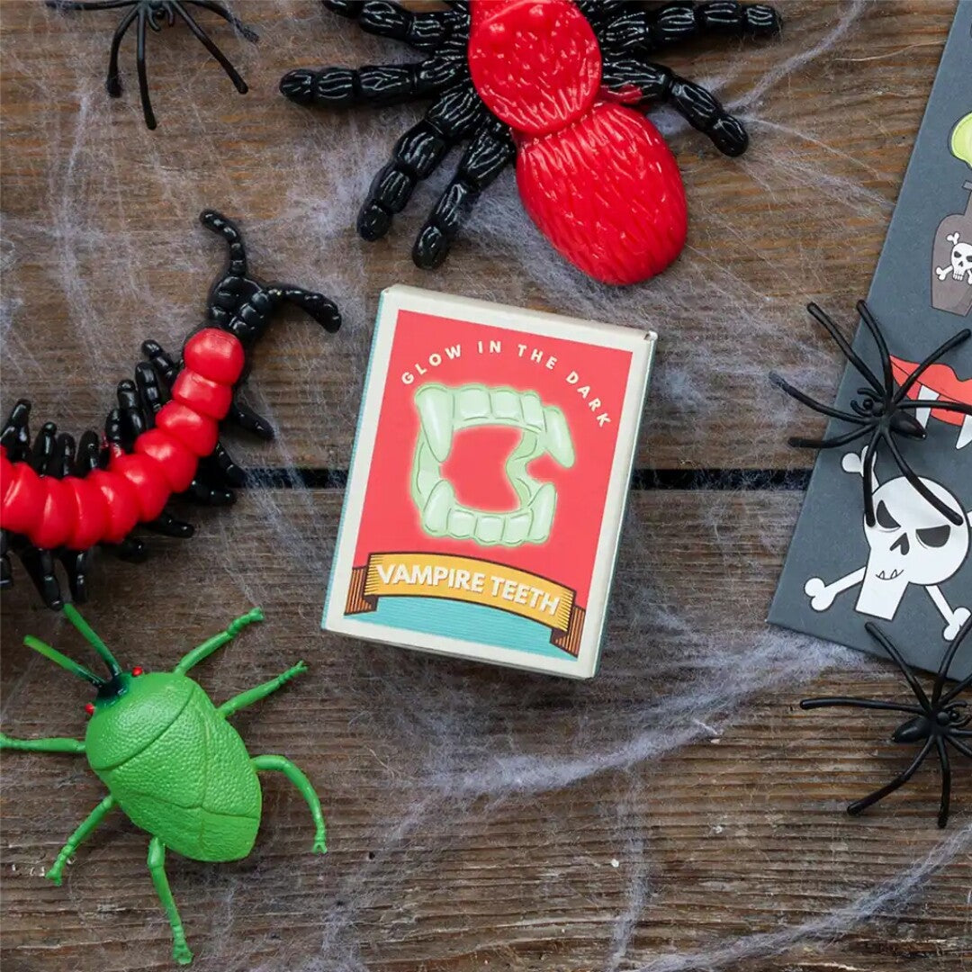 Rex London glow-in-the-dark vampiertanden verpakking op een houten ondergrond met spinnenwebben en Halloween decoraties.