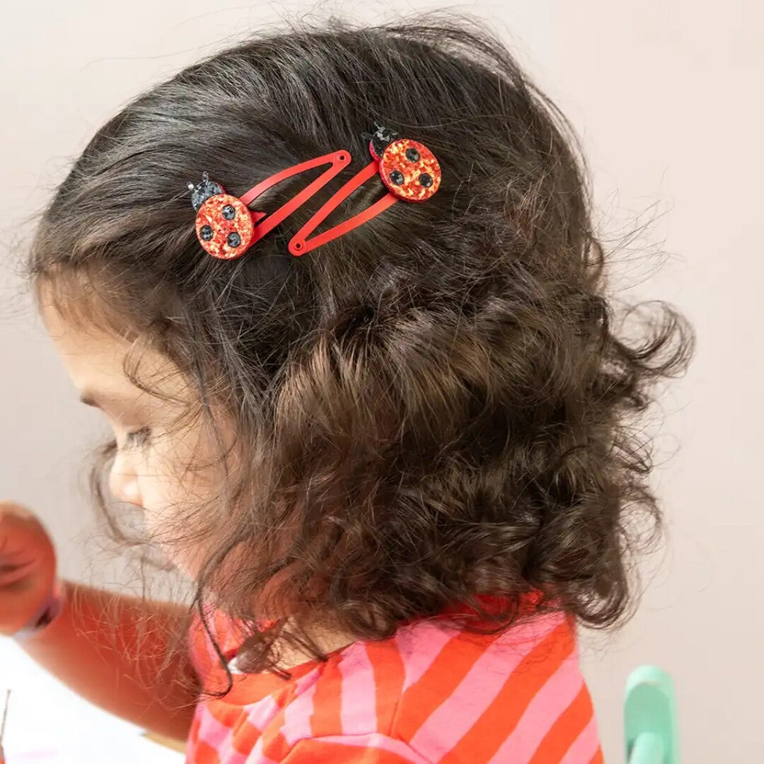 Een kind met krullend haar draagt twee rode haarspeldjes met lieveheersbeestjes.