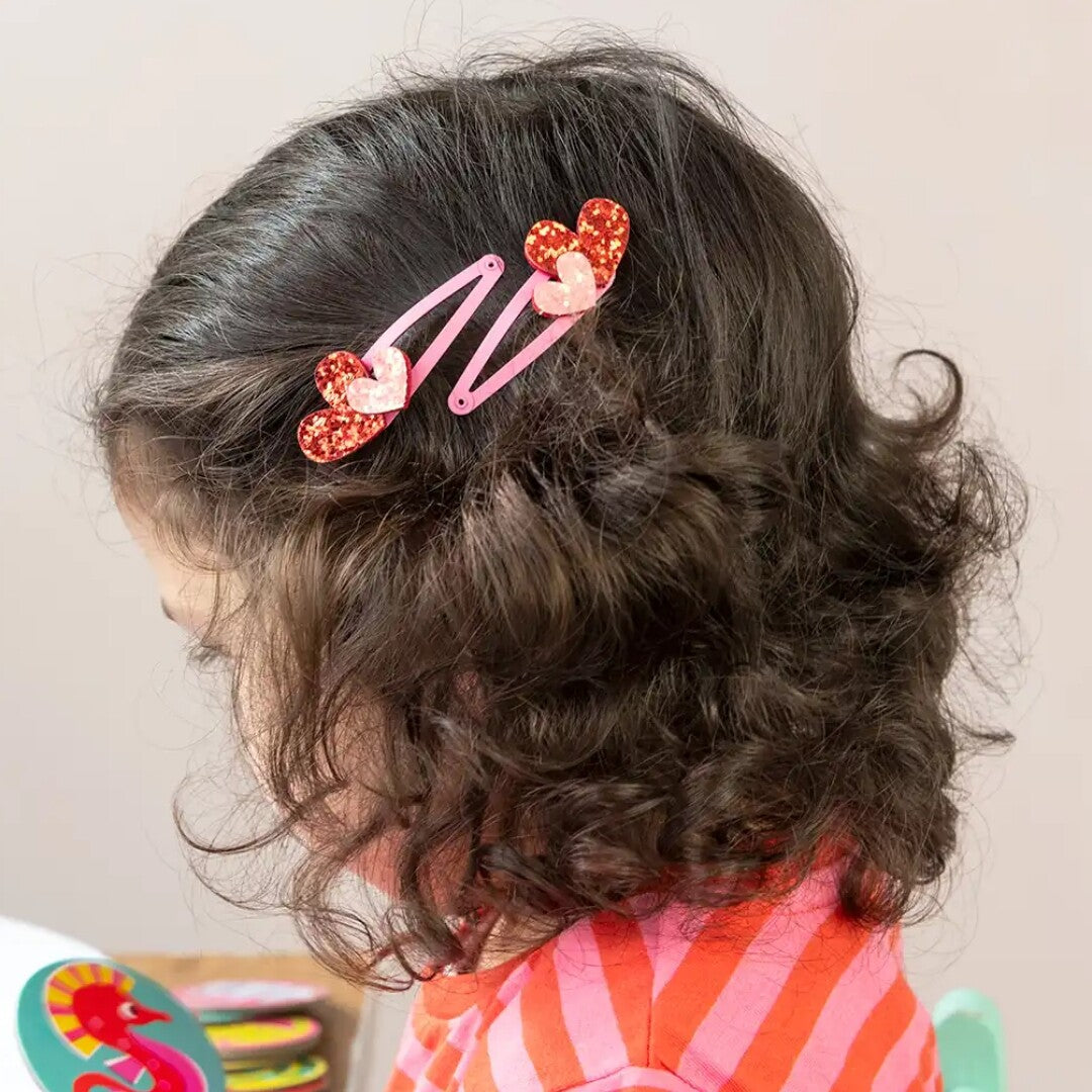 Een kind met donker krullend haar draagt twee roze haarspeldjes met glitterharten.