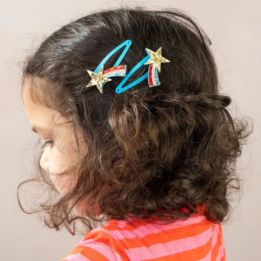 Een kind met donker krullend haar draagt de twee haarspeldjes met glitter sterren en regenboogjes.