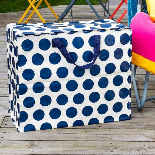 Rex London jumbo opbergtas met navy blauwe stippen op een witte achtergrond, staand op een houten terras met kleurrijke stoelen op de achtergrond.