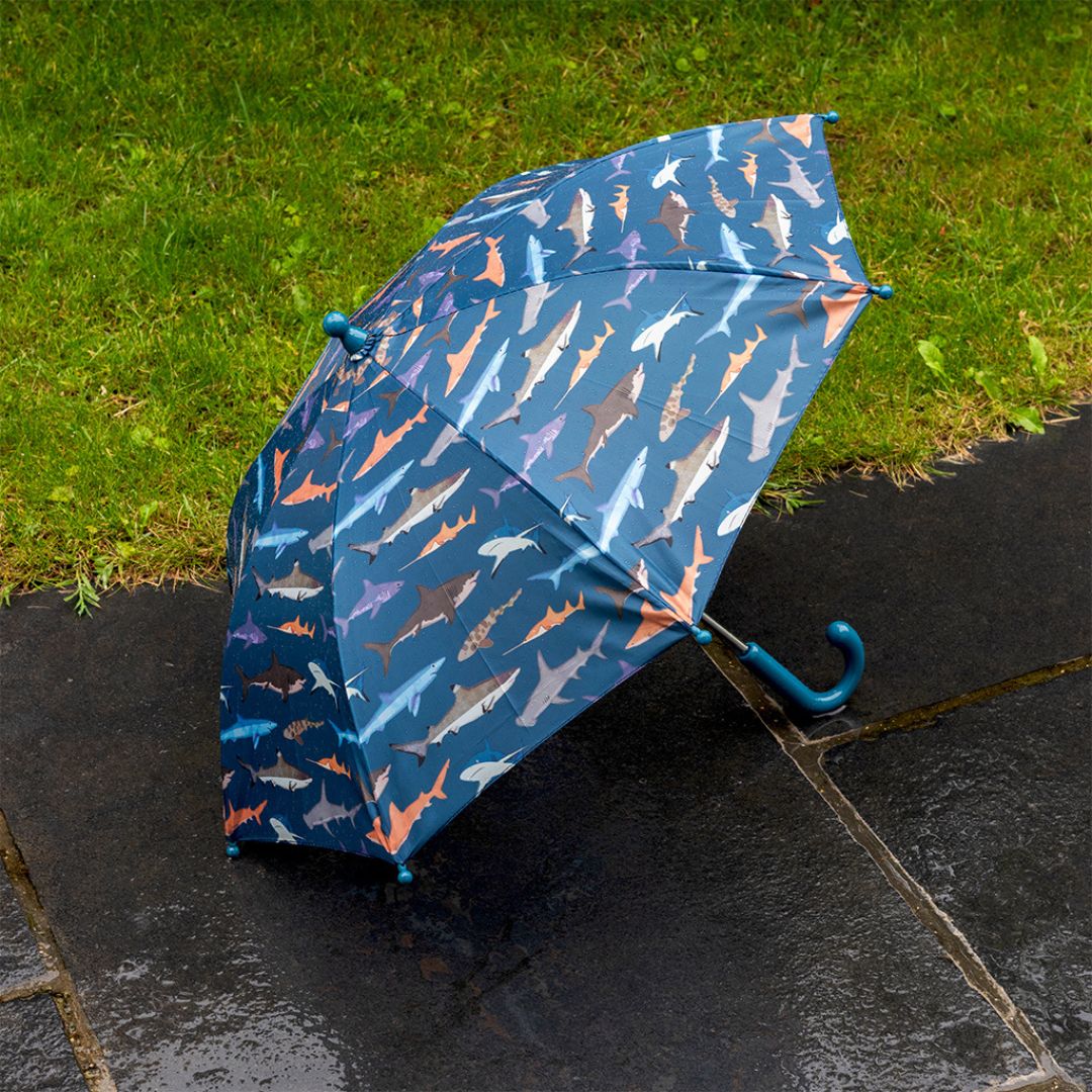 Open Rex London kinderparaplu met haaienpatroon ligt op natte tegels naast gras.