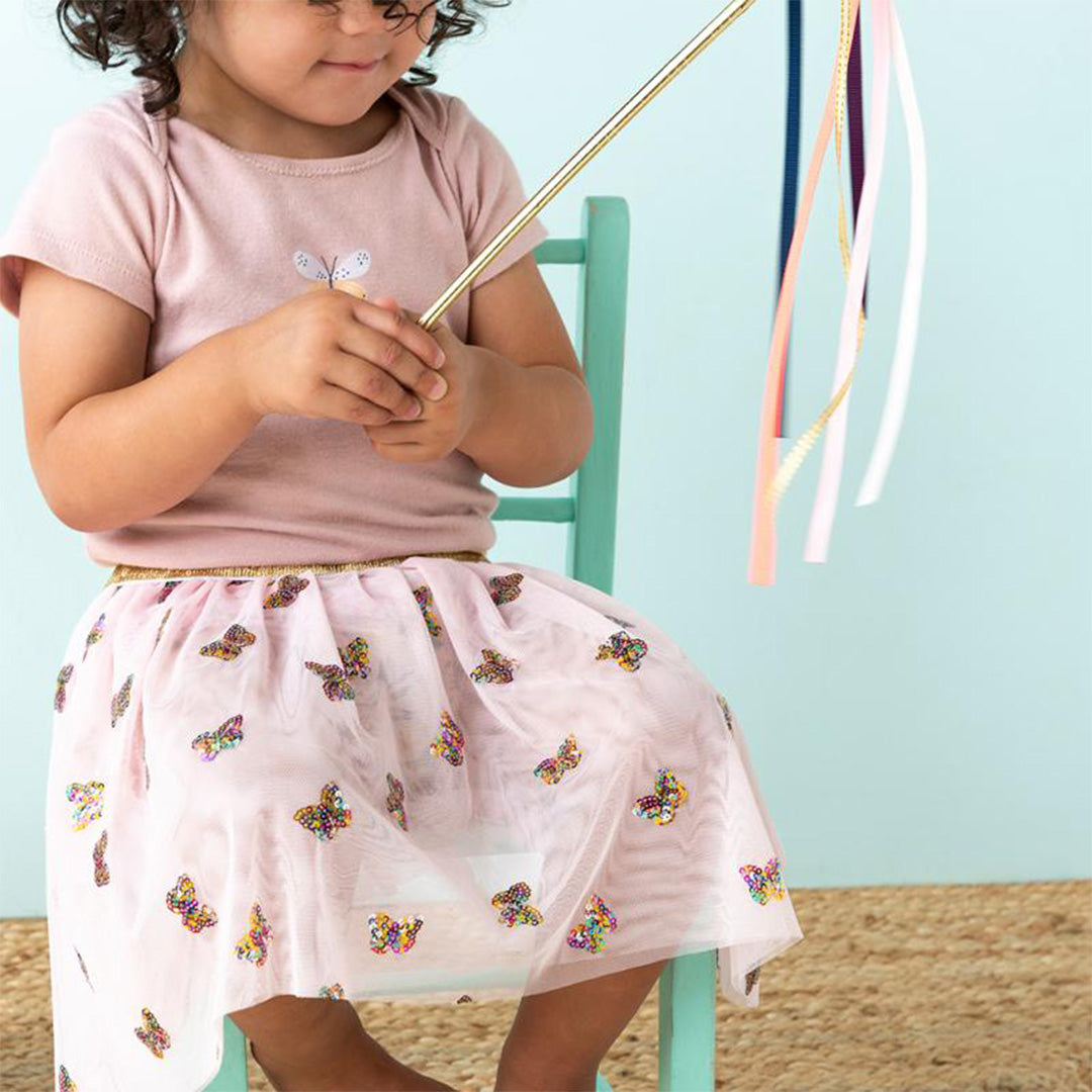 Een kind draagt de roze kinderrok met vlinderpailletten en houdt een gouden toverstaf met linten vast.