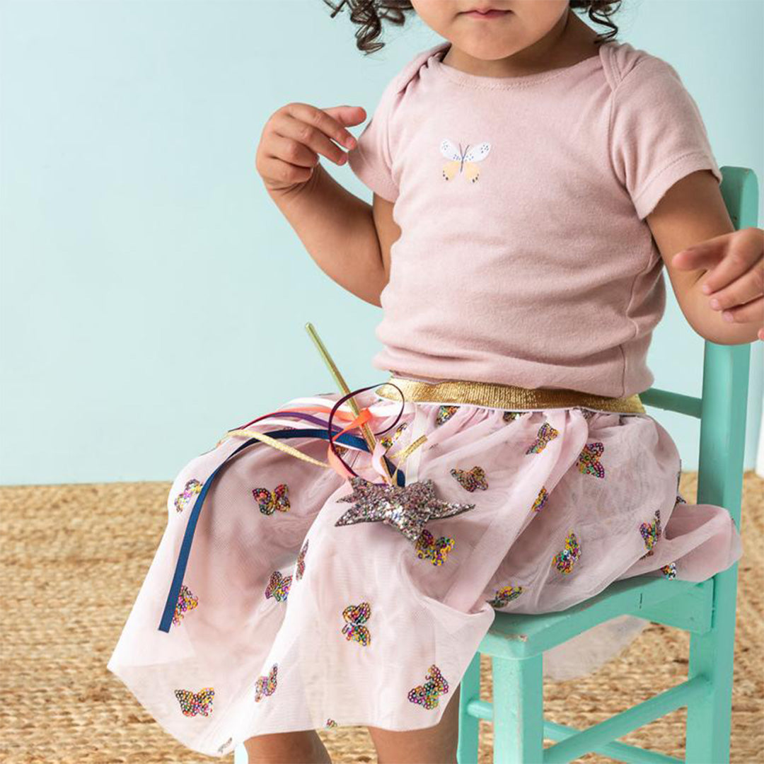 Een kind draagt de roze kinderrok met vlinderpailletten en houdt een zilveren ster-toverstaf met linten vast.