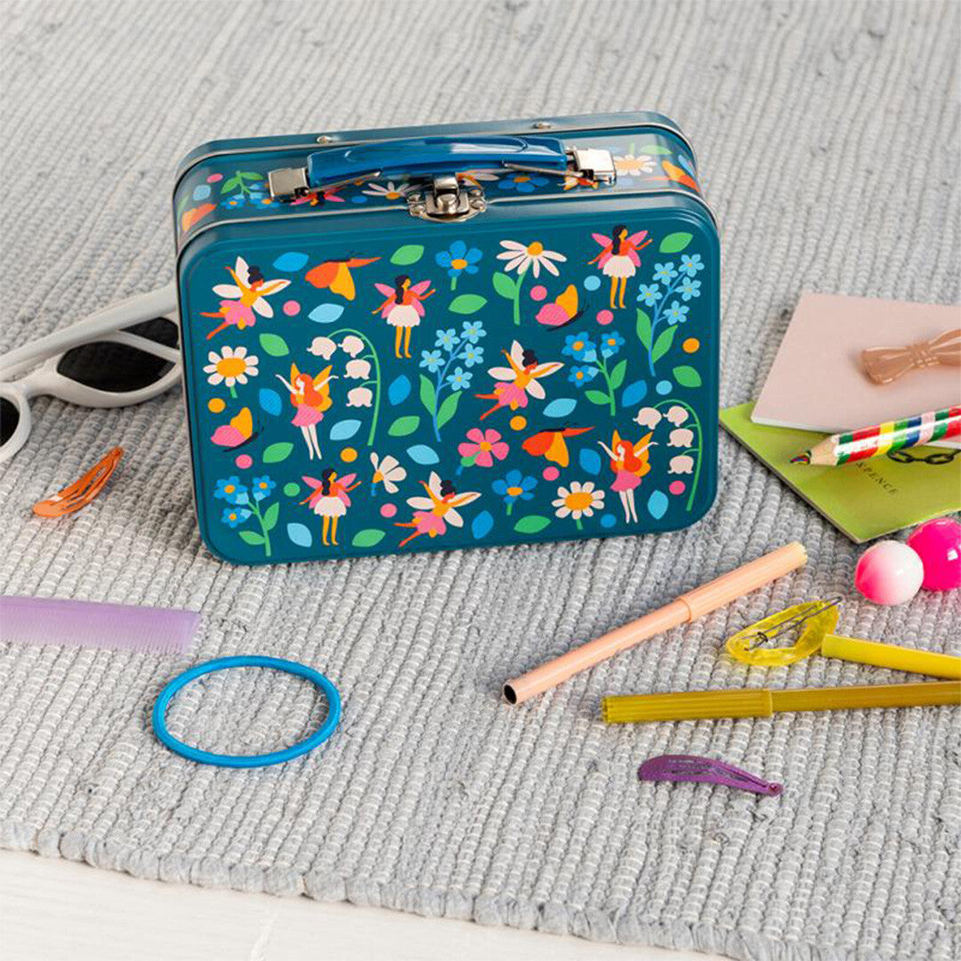 Rex London blauw metalen kinderkoffertje met feeën- en bloemenpatroon, omringd door speelgoed en schoolspullen op een grijze ondergrond.