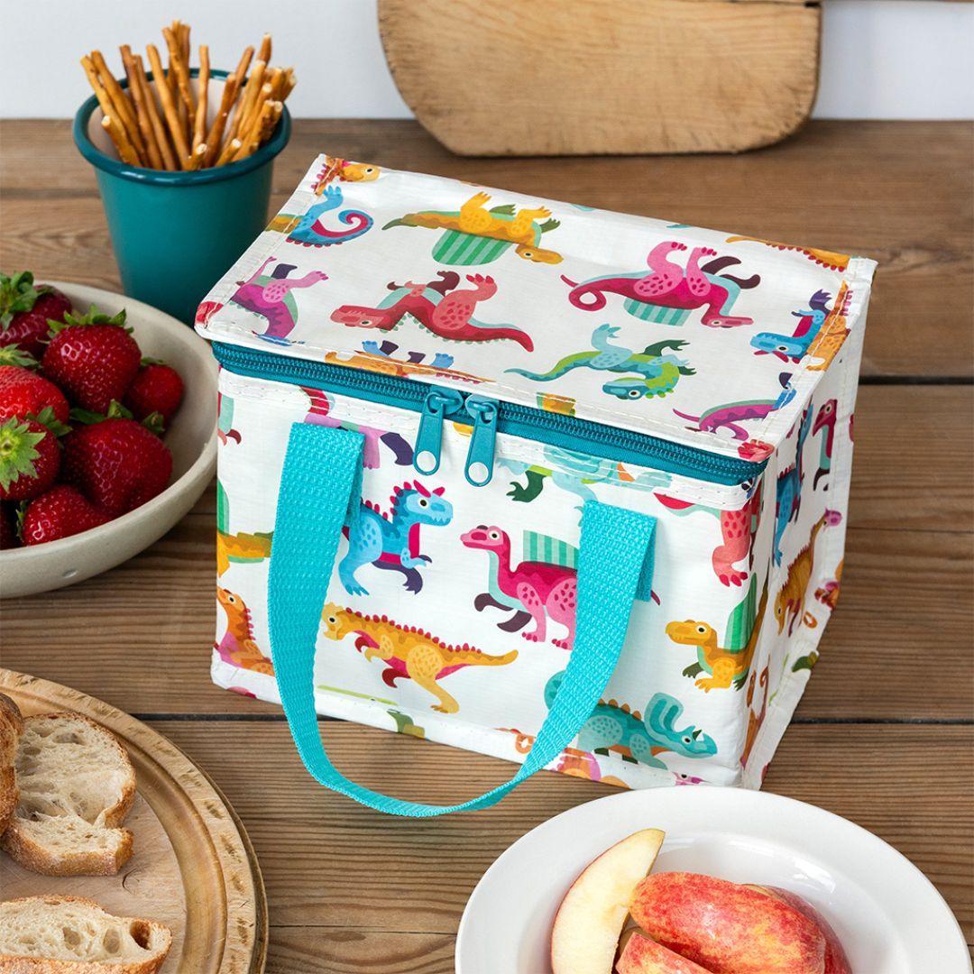 Rex London kinderlunchtas met dinosauruspatroon op een houten tafel met fruit en snacks.