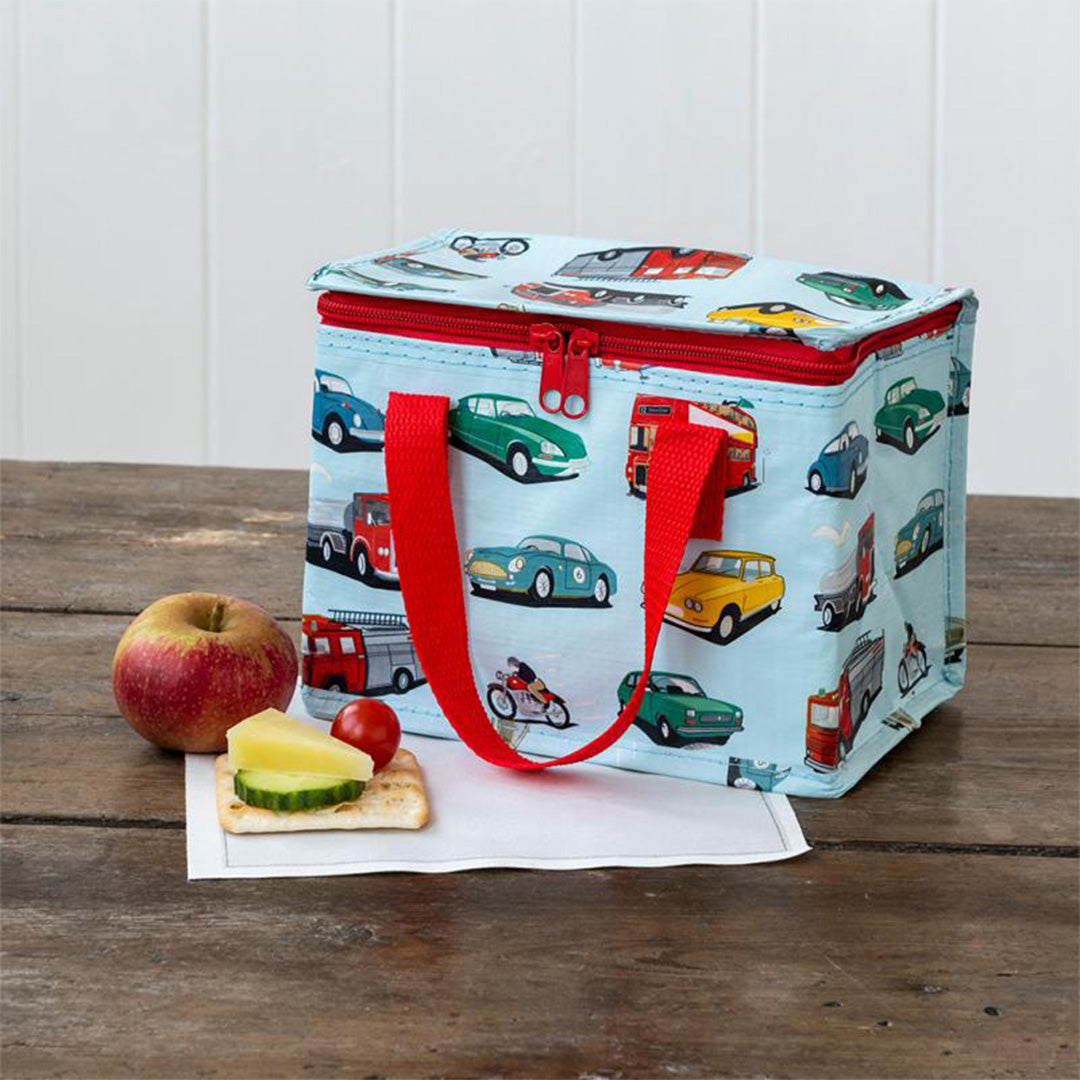 Lunchtas met voertuigpatroon op een houten tafel naast een appel en een cracker met kaas.