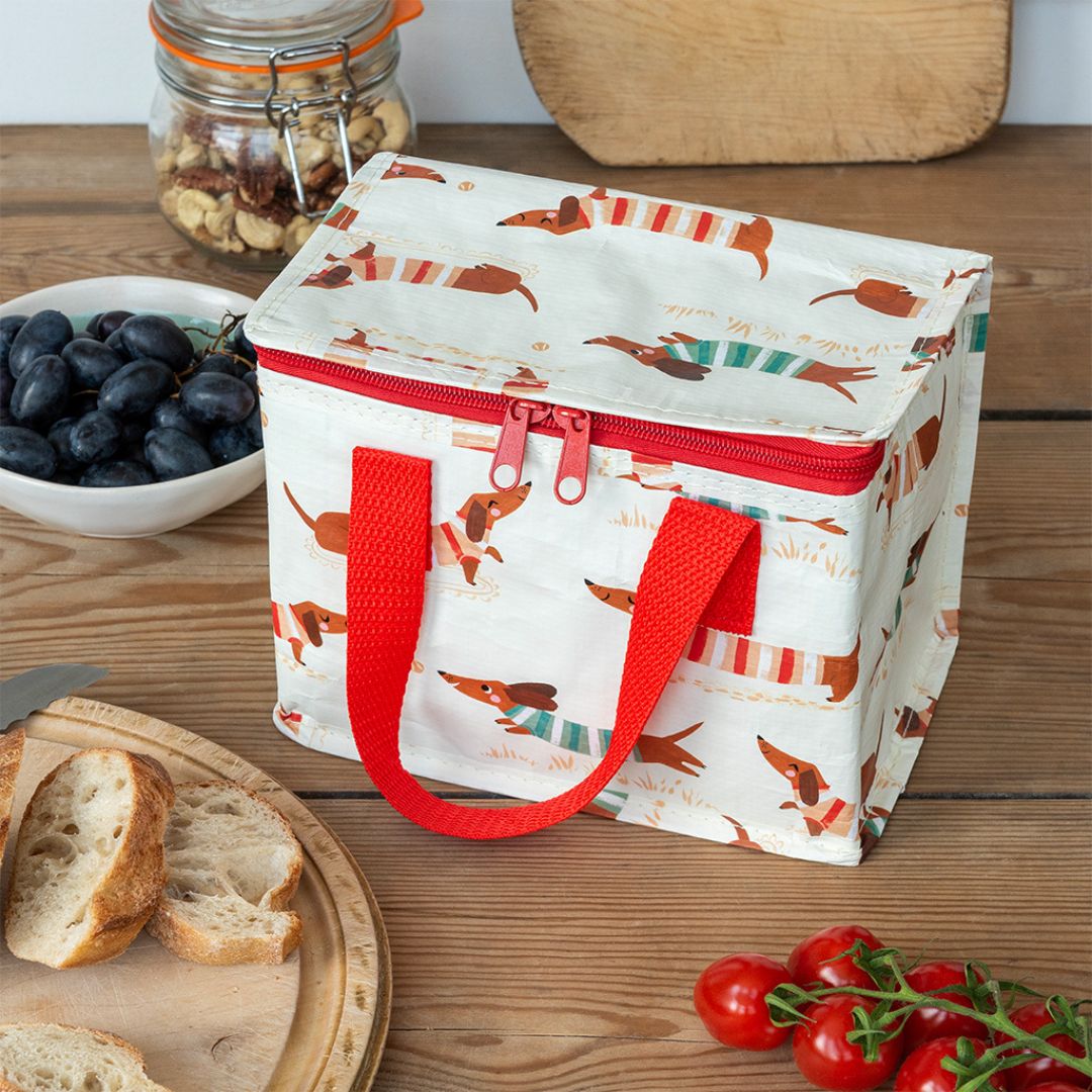 Een Rex London kinder lunchtas met teckelpatroon staat op een houten tafel tussen brood, druiven en tomaten.