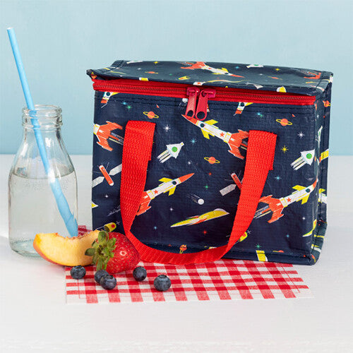 Rex London marineblauwe lunchtas voor kinderen met rakettenpatroon, staand op een tafel met een flesje drinken en fruit.