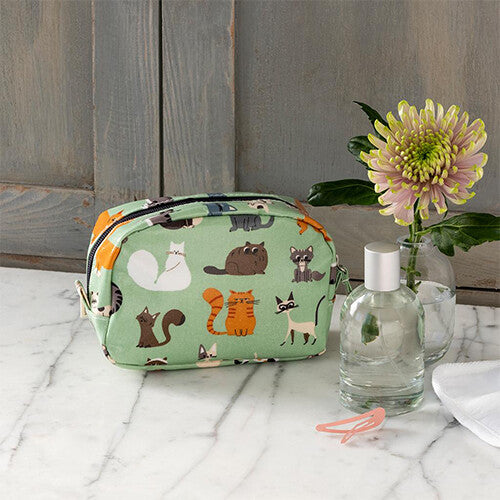 Groen make-up tasje met kattenpatroon op een marmeren ondergrond naast een parfumflesje en bloem.