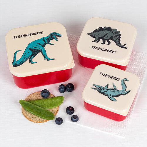 Drie Rex London snackdoosjes met dinosaurussen, gevuld met fruit en groenten op een witte ondergrond.