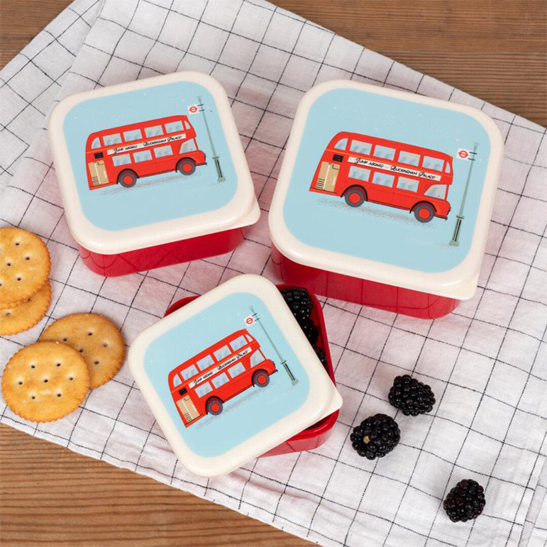 Drie rex london snackdoosjes met Londense bus illustraties, waarvan één open met bramen, naast crackers en bramen op een geruite doek.