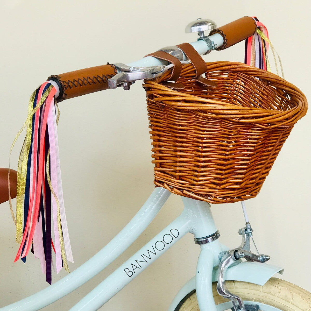 Veelkleurige Rex London stuurslingers bevestigd aan het stuur van een lichtblauwe kinderfiets met rieten mand.