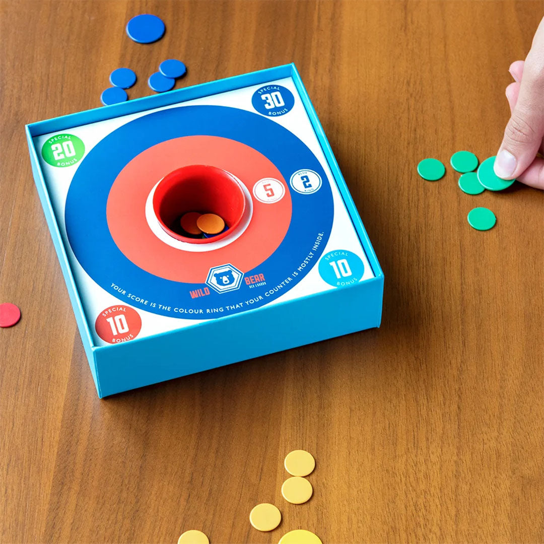 Een hand speelt het Rex London schijfjesspel op een houten tafel, met gekleurde schijfjes rondom het doelwit.