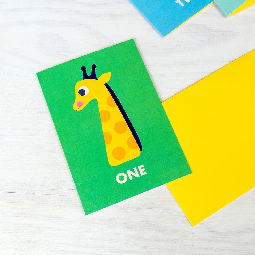 Verjaardagskaart met giraffe en het cijfer 1, liggend op een houten tafel met een gele envelop.