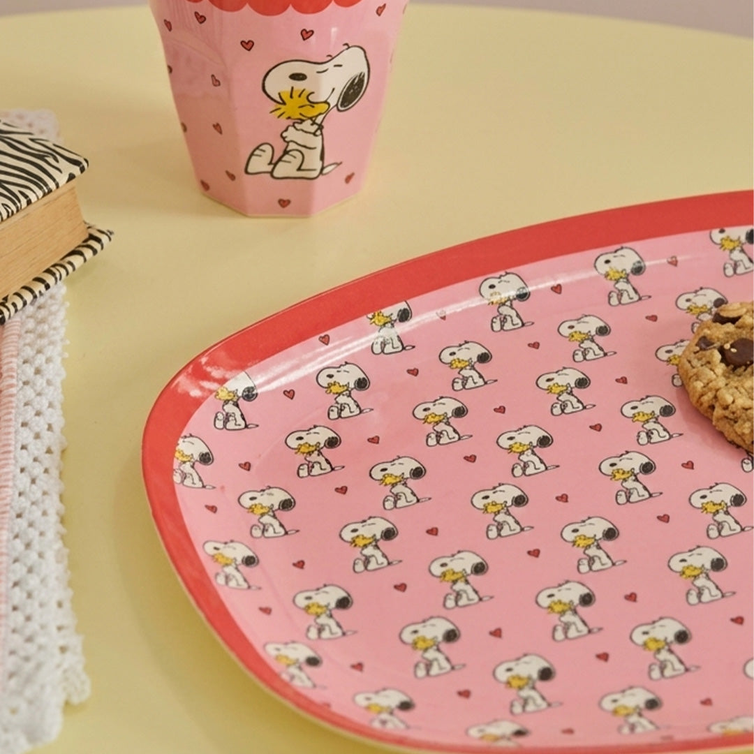 Ovaal kinderbord met Snoopy en harten, samen met een bijpassende beker en een koekje op een gele ondergrond.