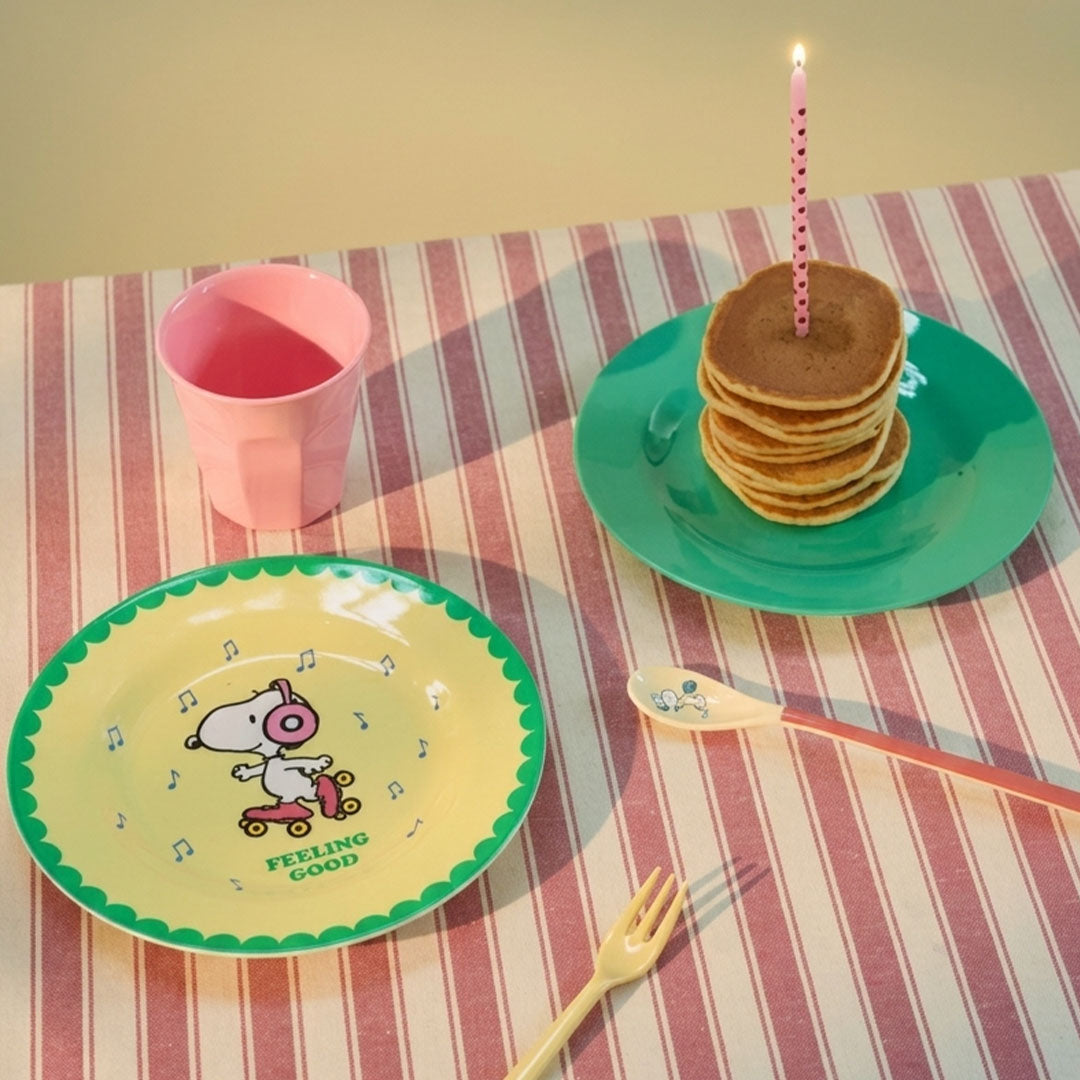 Een tafelsetting met het rice kinderbord met Snoopy, een roze beker, een groen bord met pannenkoeken en een kaars.