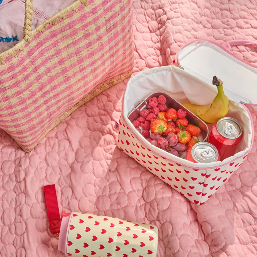 Open Rice koeltas met rode harten, gevuld met fruit en blikjes, op een roze deken.