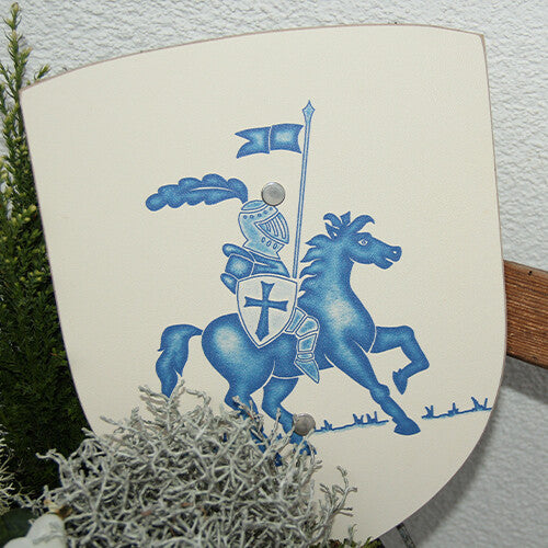 Vah houten speelgoedschild met blauwe ridder, staand tussen groene planten.