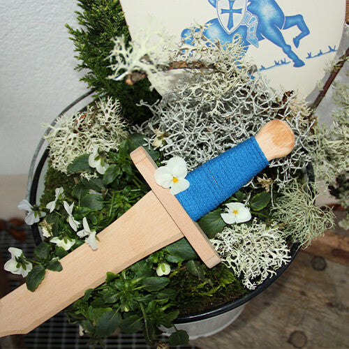 Vah houten speelgoedzwaard met blauw handvat, liggend in een pot met planten en witte bloemen.