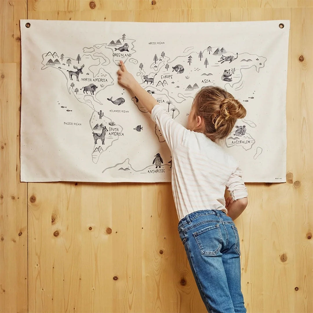 Een meisje wijst naar Groenland op het rose in April wandkleed met wereldkaart en dierenillustraties aan een houten muur.