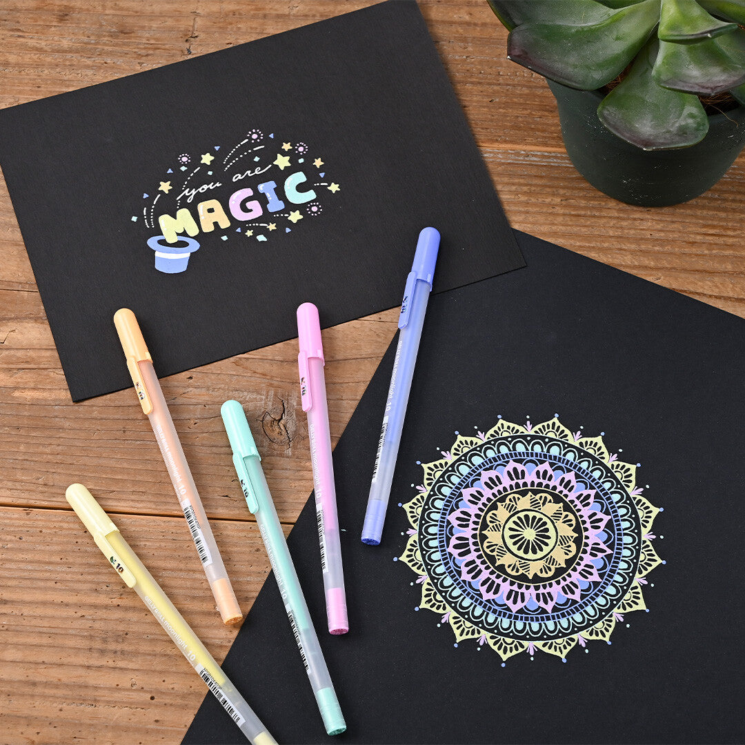 Vijf Sakura Gelly Roll Moonlight gelpennen liggen op een houten tafel naast twee zwarte vellen papier met kleurrijke tekeningen.