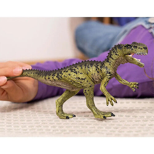 Schleich plastic dinosaurusfiguur Monolophosaurus op een ondergrond, vastgehouden bij de staart door een hand.