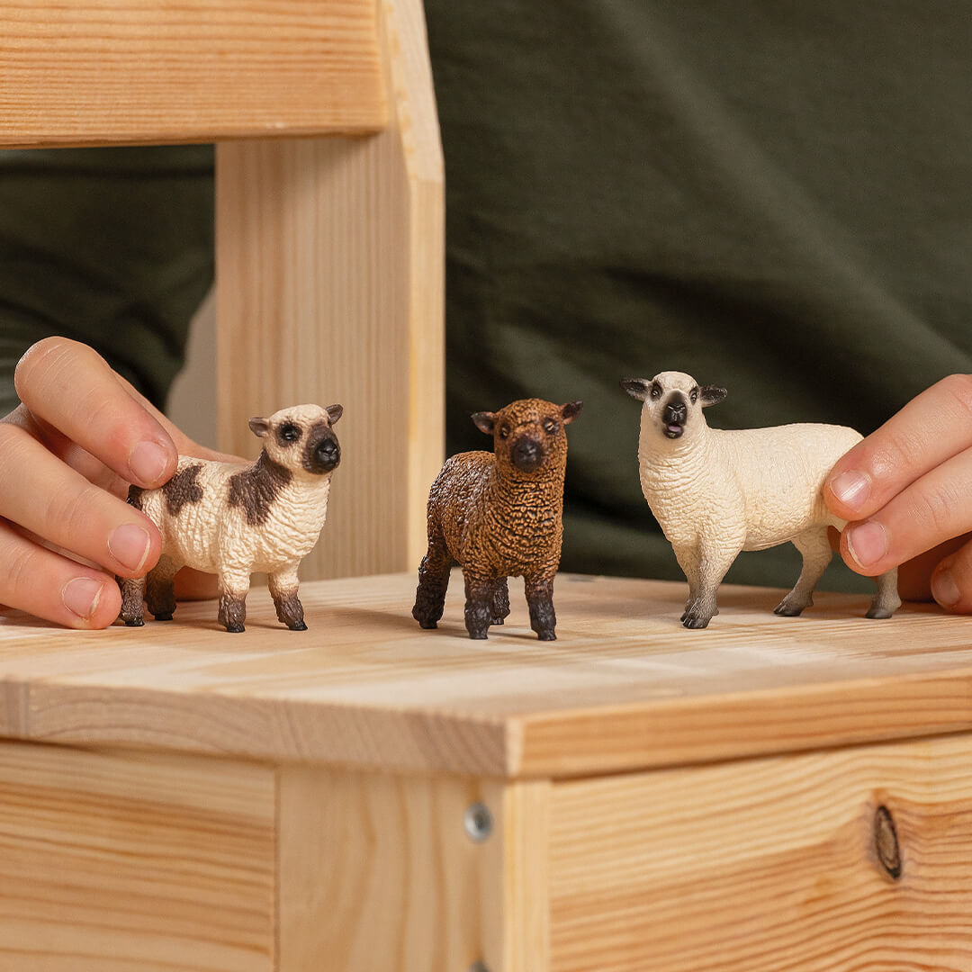Schleich dierenfiguurtjes van drie schapen en lammetjes op een houten stoel, vastgehouden door kinderhanden.