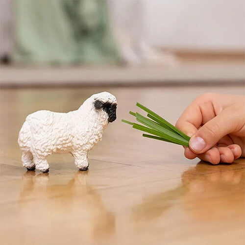 Het Schleich Walliser zwartneusschaap dierenfiguurtje staat op een houten vloer en kijkt naar een hand met gras.