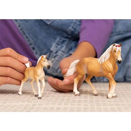 Twee Schleich paardenfiguurtjes, een Haflinger veulen en een volwassen paard, worden vastgehouden door kinderhanden.