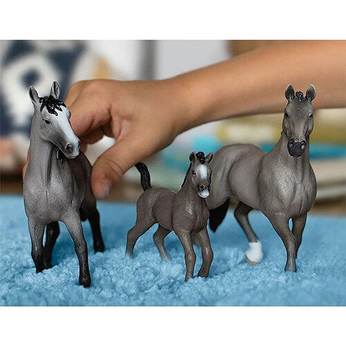 Drie Schleich paarden dierenfiguurtjes, waaronder een merrie en een veulen, op een blauwe ondergrond met een kinderhand op de achtergrond.